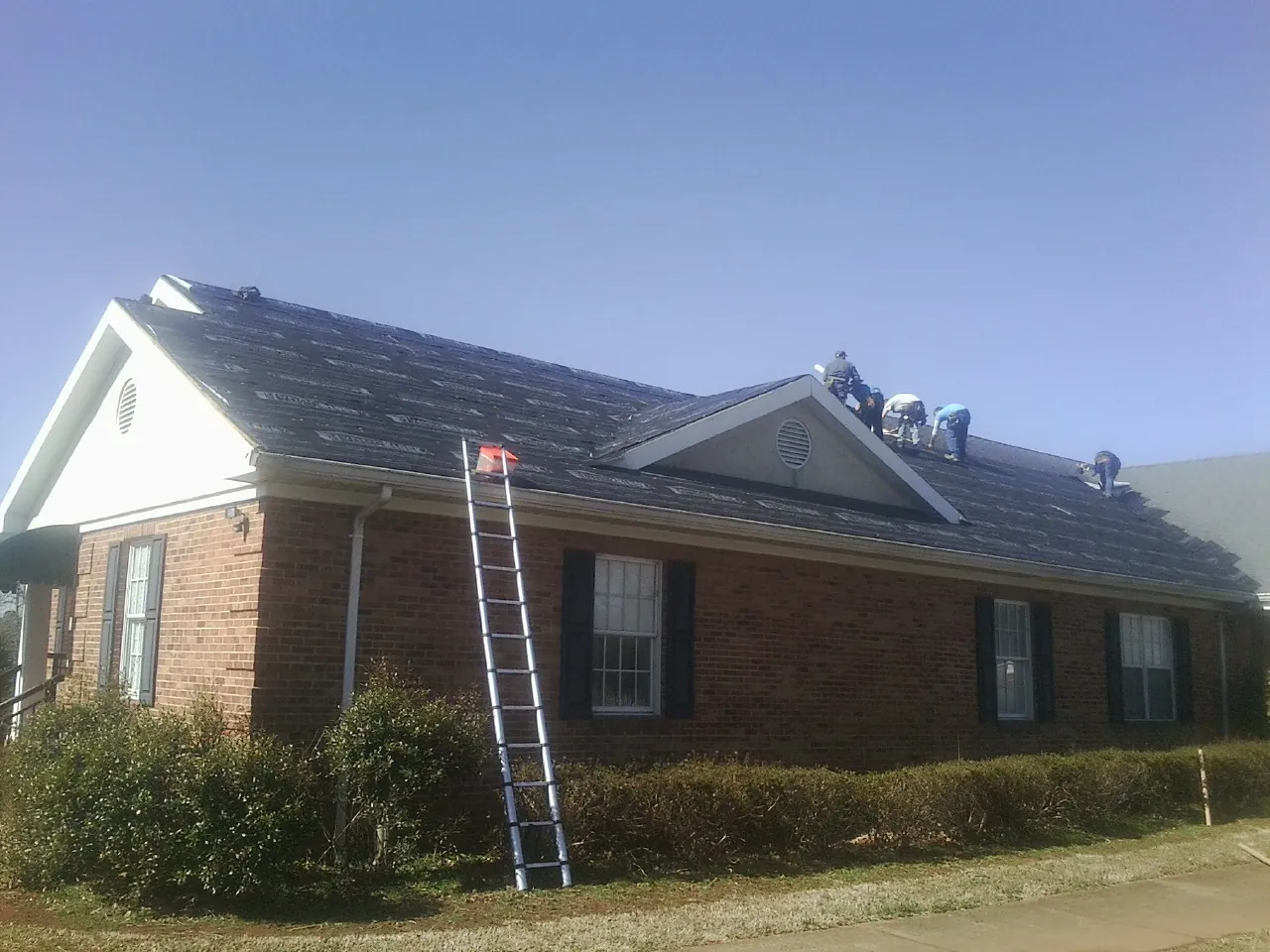 Crew tearing shingles off a brick ranch roof with a ladder set — Metal Roof Repair work by our crew in Ray City
